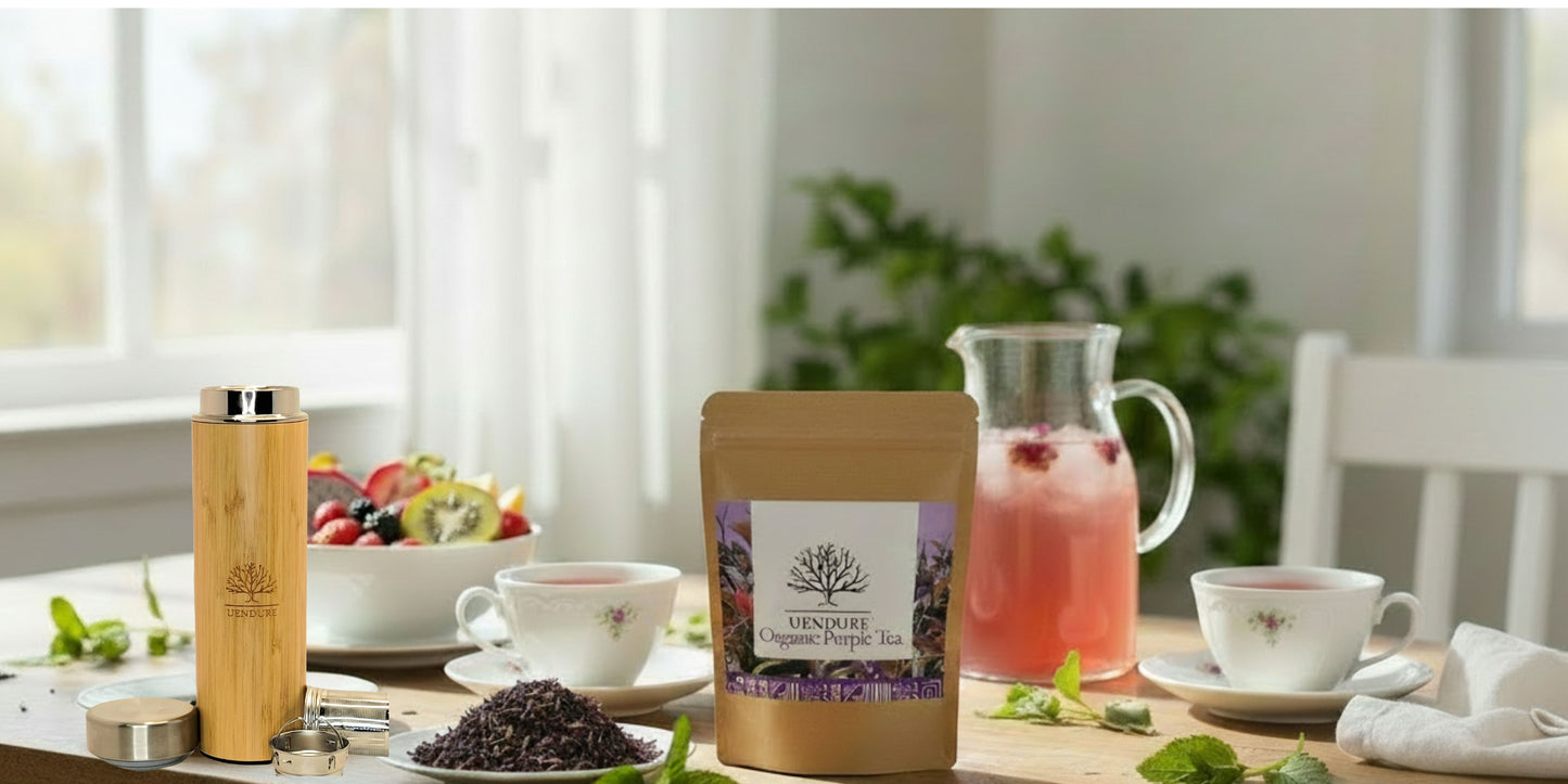 A stand-up pouch of lavender herbal tea stands on a dining table, surrounded by cups, a pitcher of pink drink, a bamboo travel tumbler, and fruit in a bright indoor setting.