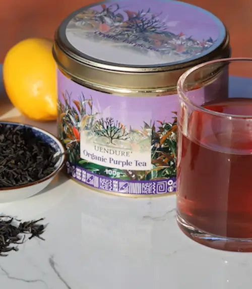 A tin of Undure Organic Purple Tea on a white marble surface, with a glass of brewed purple tea, loose tea leaves, and a lemon in the background.