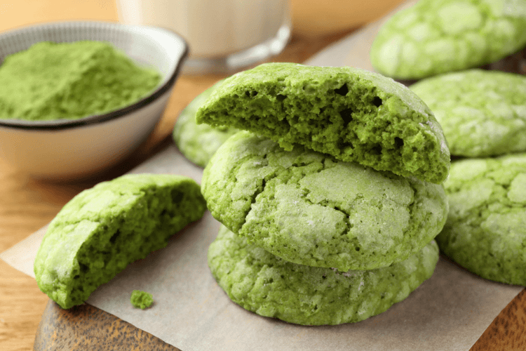 
                    
                      Green matcha cookies on a wooden surface with a bowl of blueberry matcha powder in the background.
                    
                  